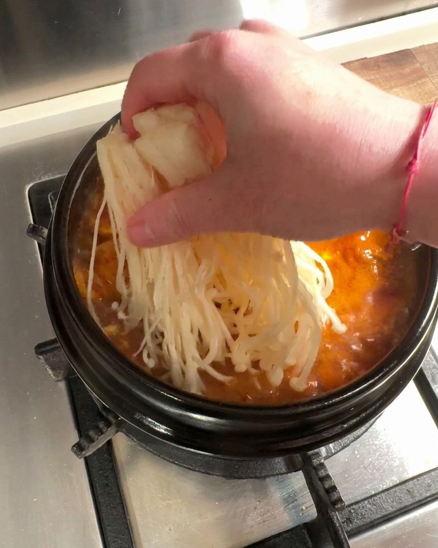 Champignons enoki frais et tranches de bœuf cru placés dans le ragoût au kimchi rouge bouillonnant.