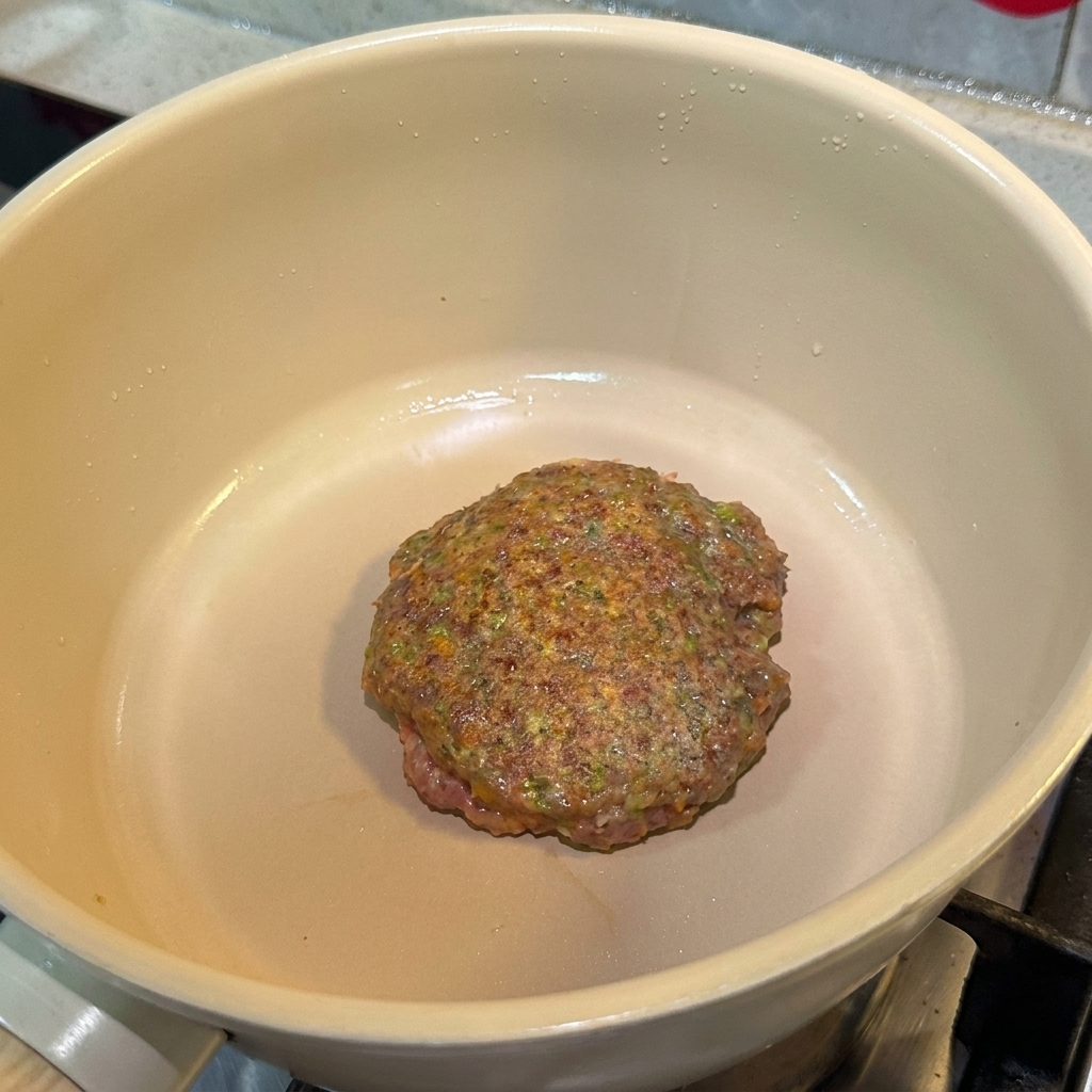 Une galette de bœuf assaisonnée en train de cuire dans une poêle, montrant une surface bien dorée.