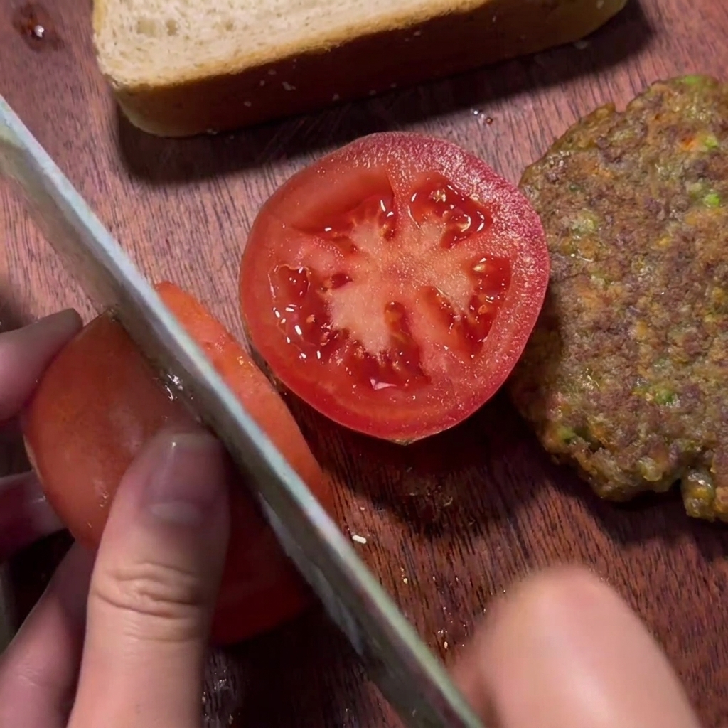 Une main tranchant une tomate rouge fraîche sur une planche en bois à côté dune galette de bœuf cuite.