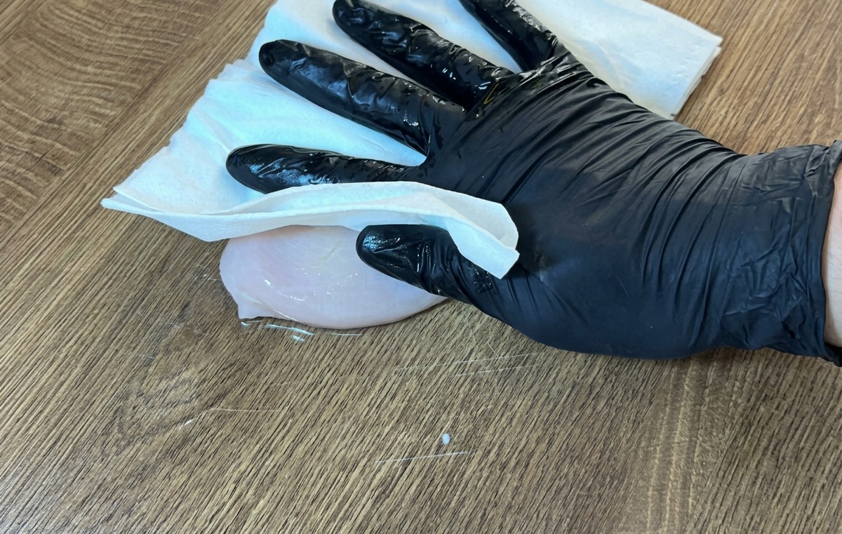 Une main gantée utilisant du papier absorbant pour sécher un blanc de poulet cru sur une surface en bois.