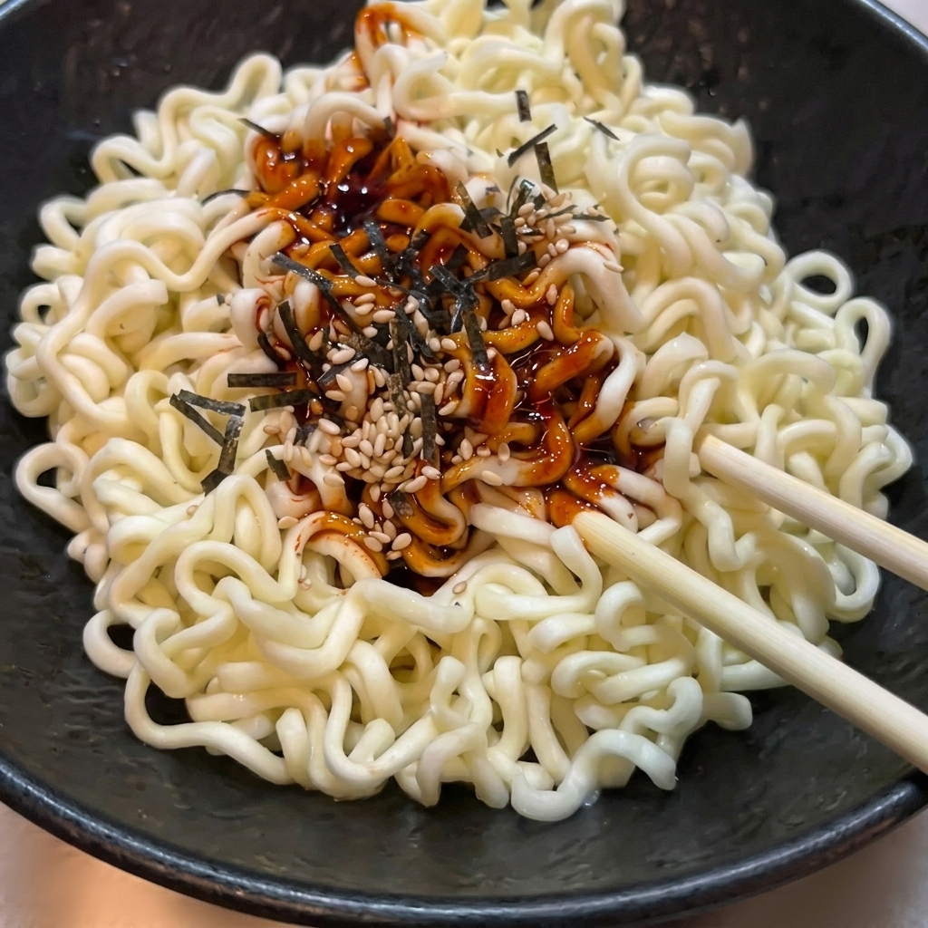 Nouilles ramen ondulées dans un bol noir texturé, mélangées avec une sauce rouge épicée, des graines de sésame et des algues.