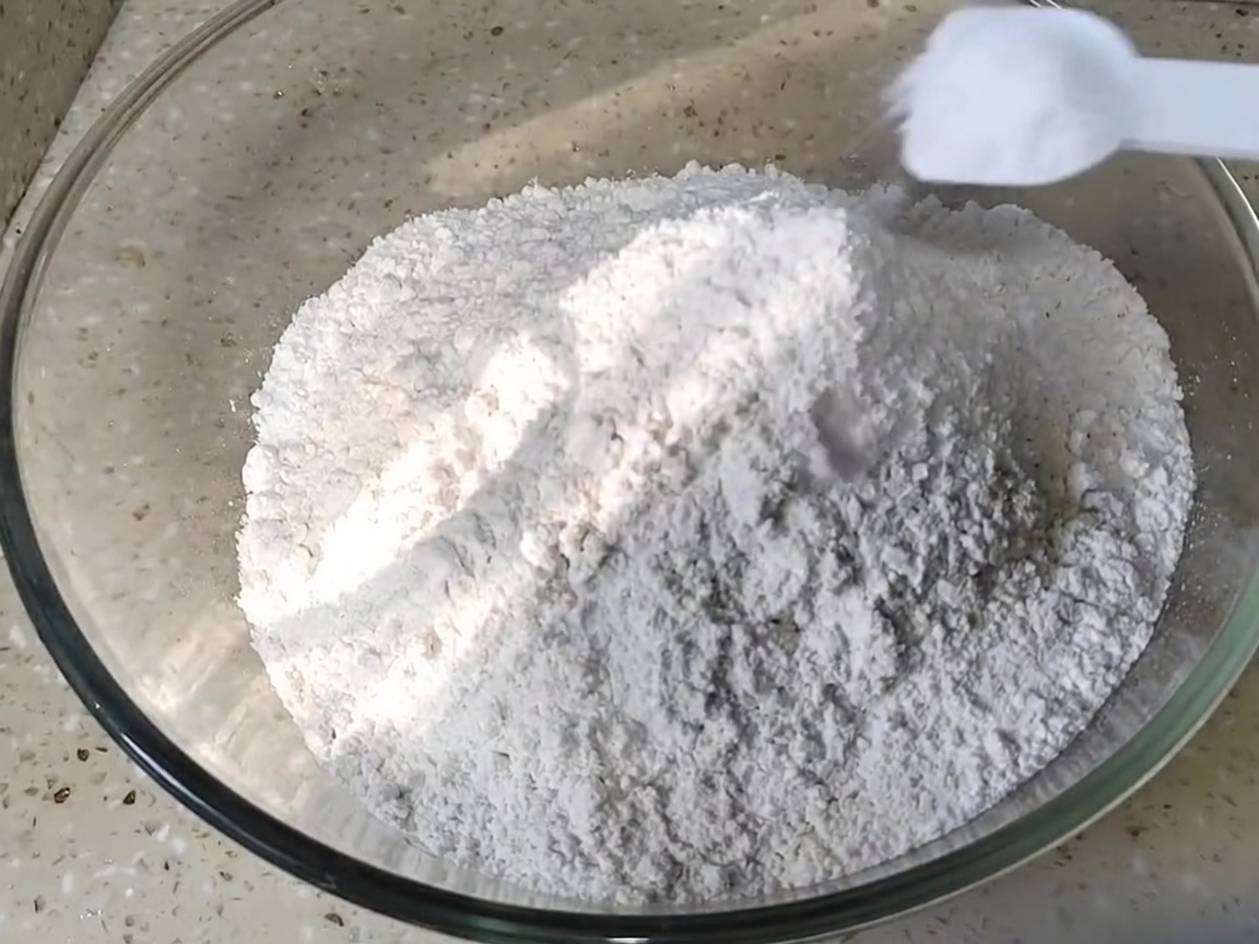 Une cuillerée de sel ajoutée à un tas de farine blanche dans un bol mélangeur en verre.