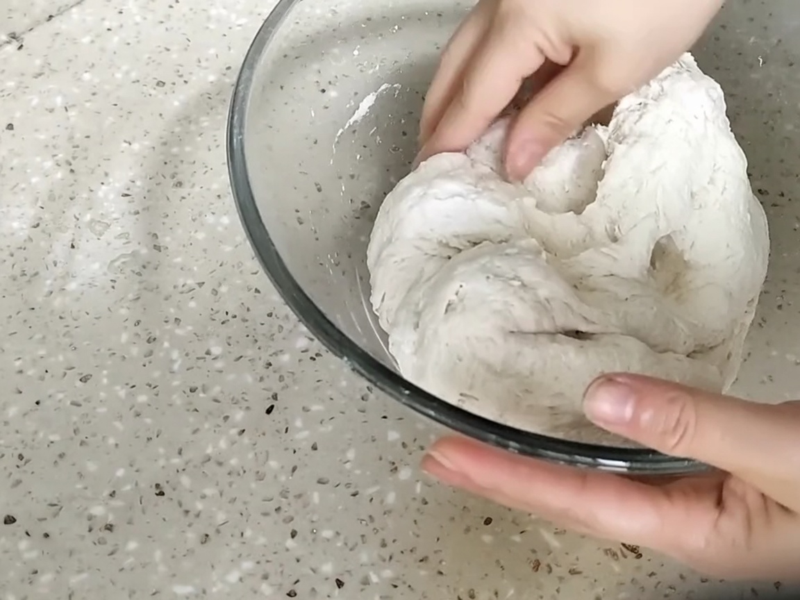 Des mains pressant et pétrissant une boule de pâte rugueuse dans un bol en verre.