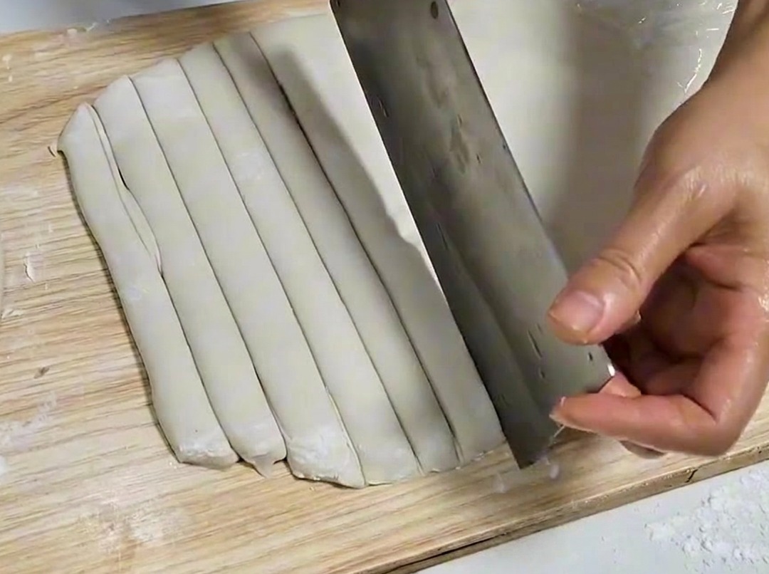 Feuille de pâte coupée en bandes égales avec un couperet en métal sur une planche en bois.