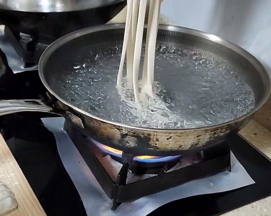 Main étirant une bande de pâte en une longue nouille avant de la déposer dans une casserole deau bouillante.