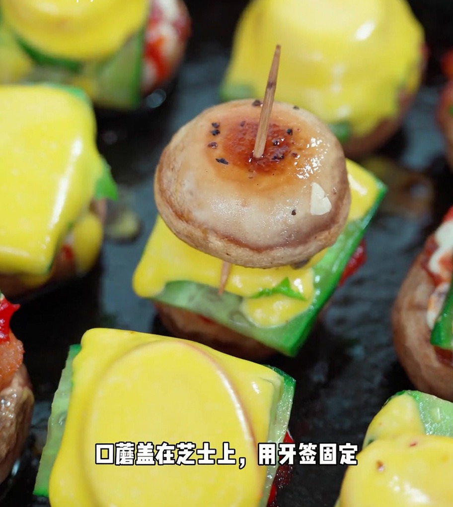 Un mini burger de champignon fini, maintenu par un cure-dent, montrant les couches empilées de champignon, concombre, laitue, tomate, fromage et un second chapeau.