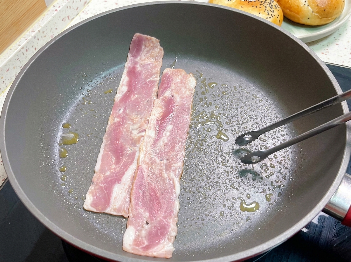 Tranches de bacon en train de frire dans une poêle chaude, retournées avec une pince.