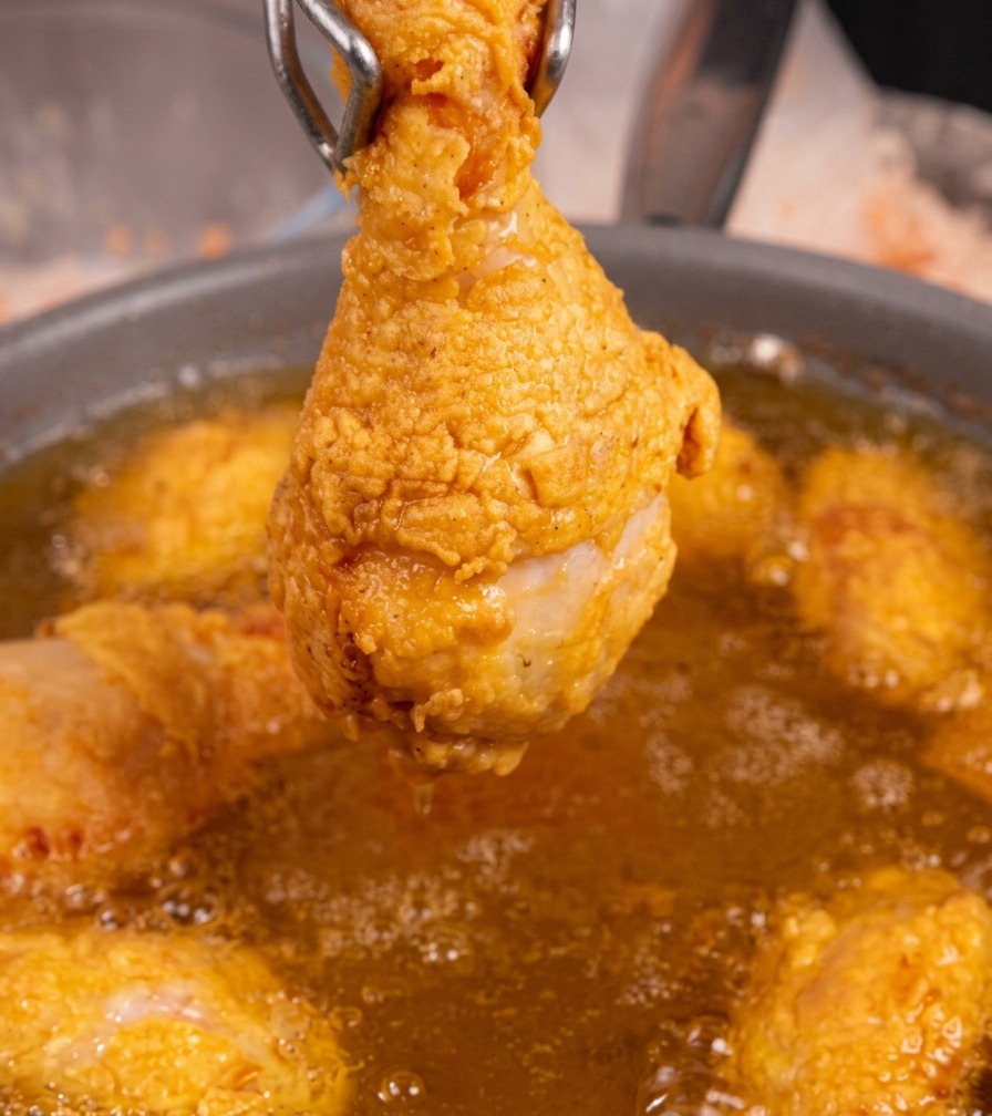Un seul pilon de poulet frit bien doré tenu par des pinces en métal au-dessus dune poêle dhuile bouillonnante.