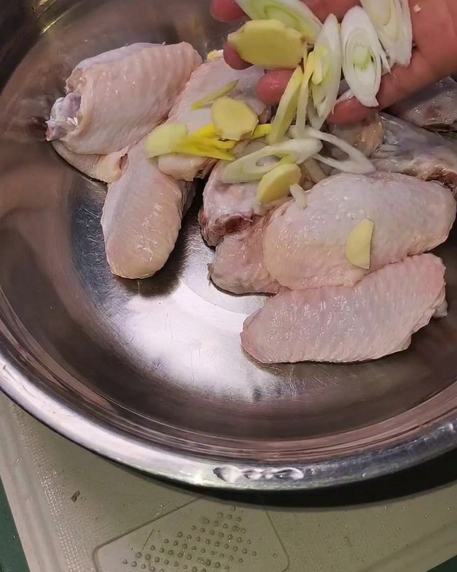 Ailes de poulet crues dans un bol en métal garnies de tranches de gingembre, dail et doignons verts prêtes à être assaisonnées.