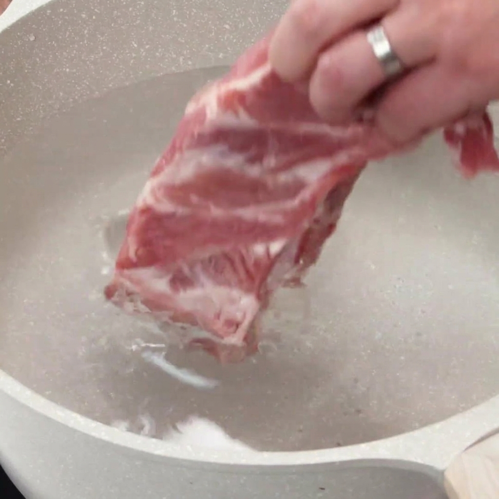 Une main plaçant un rack de travers de porc crus dans une casserole remplie deau froide.