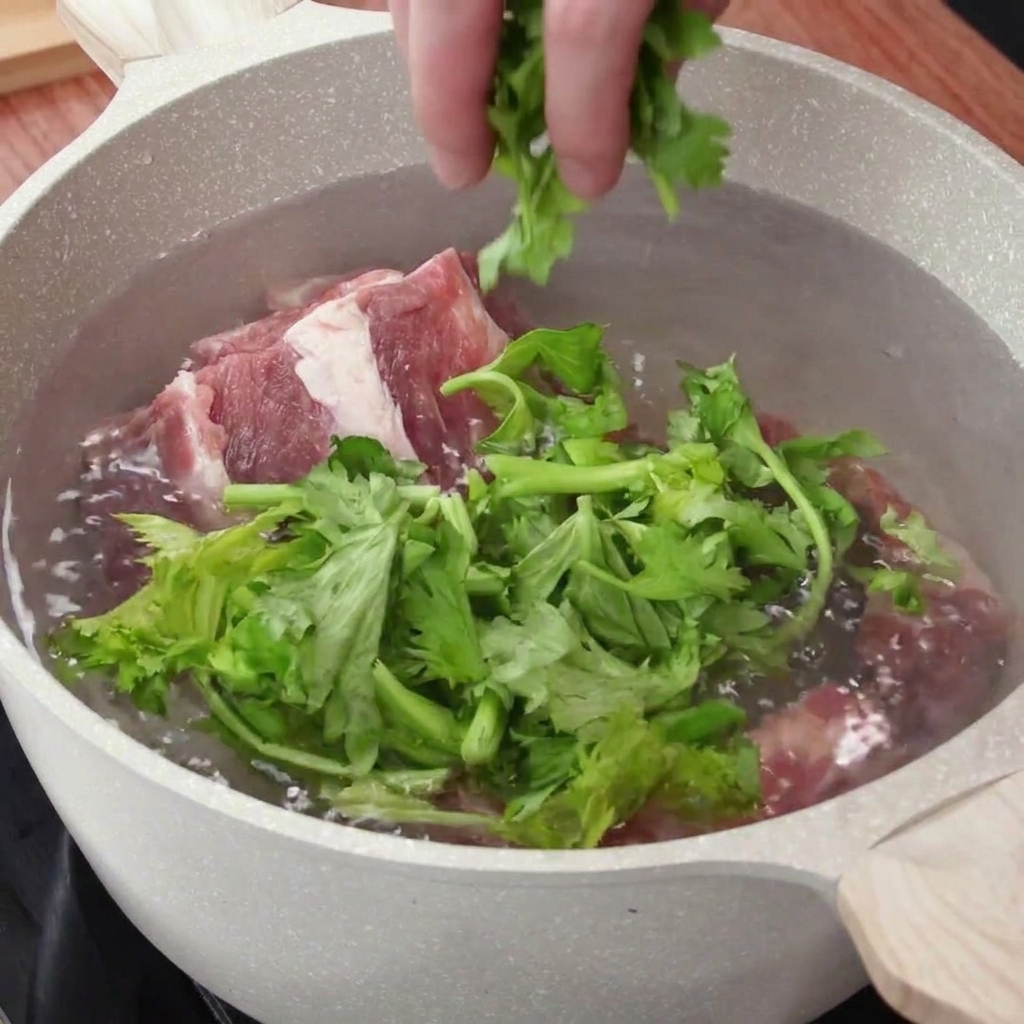Une main ajoutant des feuilles de céleri hachées et des aromates dans une casserole deau contenant des travers de porc crus.