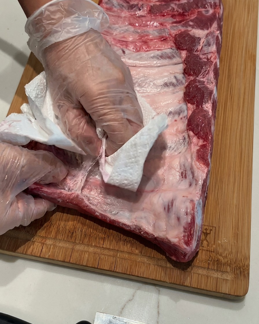 Mains portant des gants en plastique retirant une fine membrane dun rack de porc cru sur une planche en bois.