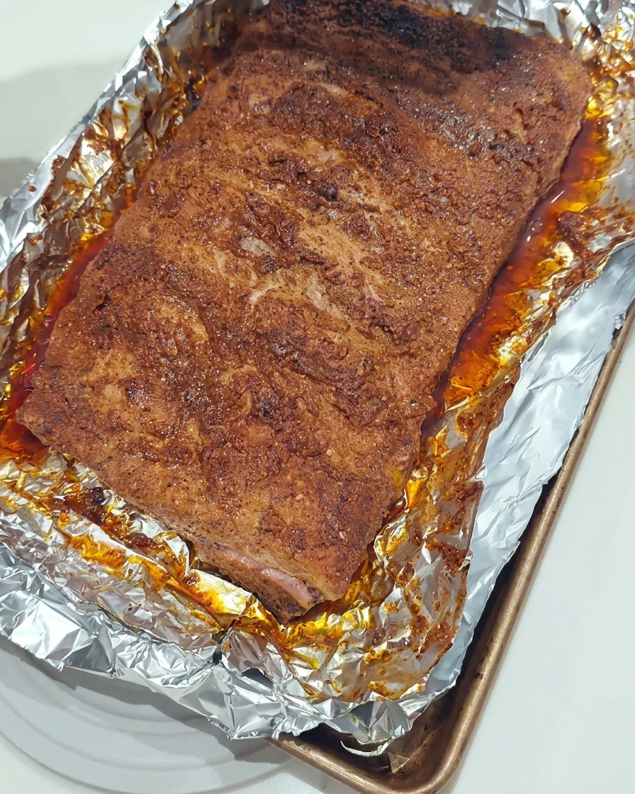 Un pinceau en silicone appliquant une couche brillante de sauce barbecue sur des travers de porc rôtis dans un plat.