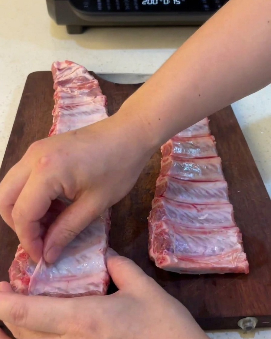 Gros plan montrant des mains retirant une fine membrane translucide au dos de travers de porc crus.