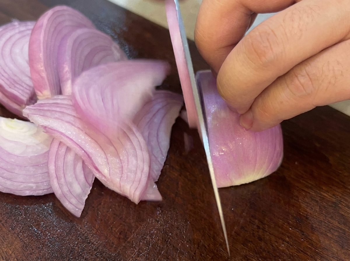 Un couteau tranchant un oignon rouge en lamelles sur une planche à découper en bois foncé.
