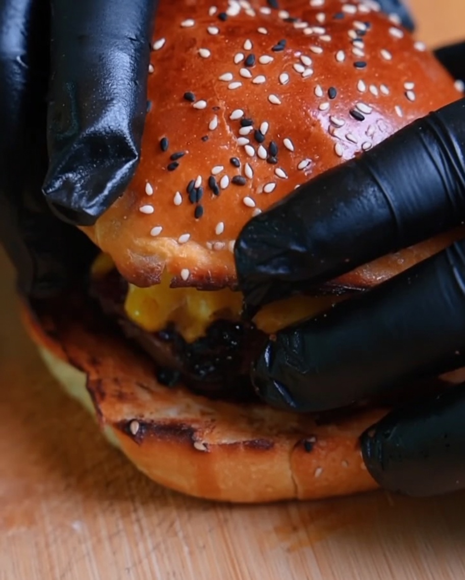 Gros plan dun burger finalisé, avec le pain supérieur ajouté par des mains gantées.