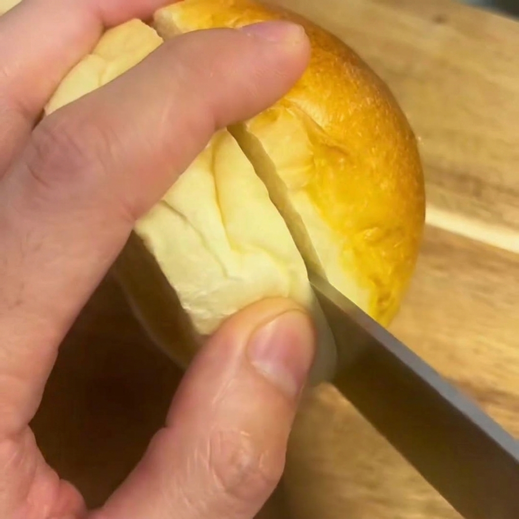 Un couteau coupant un pain à burger moelleux en deux sur une planche en bois.