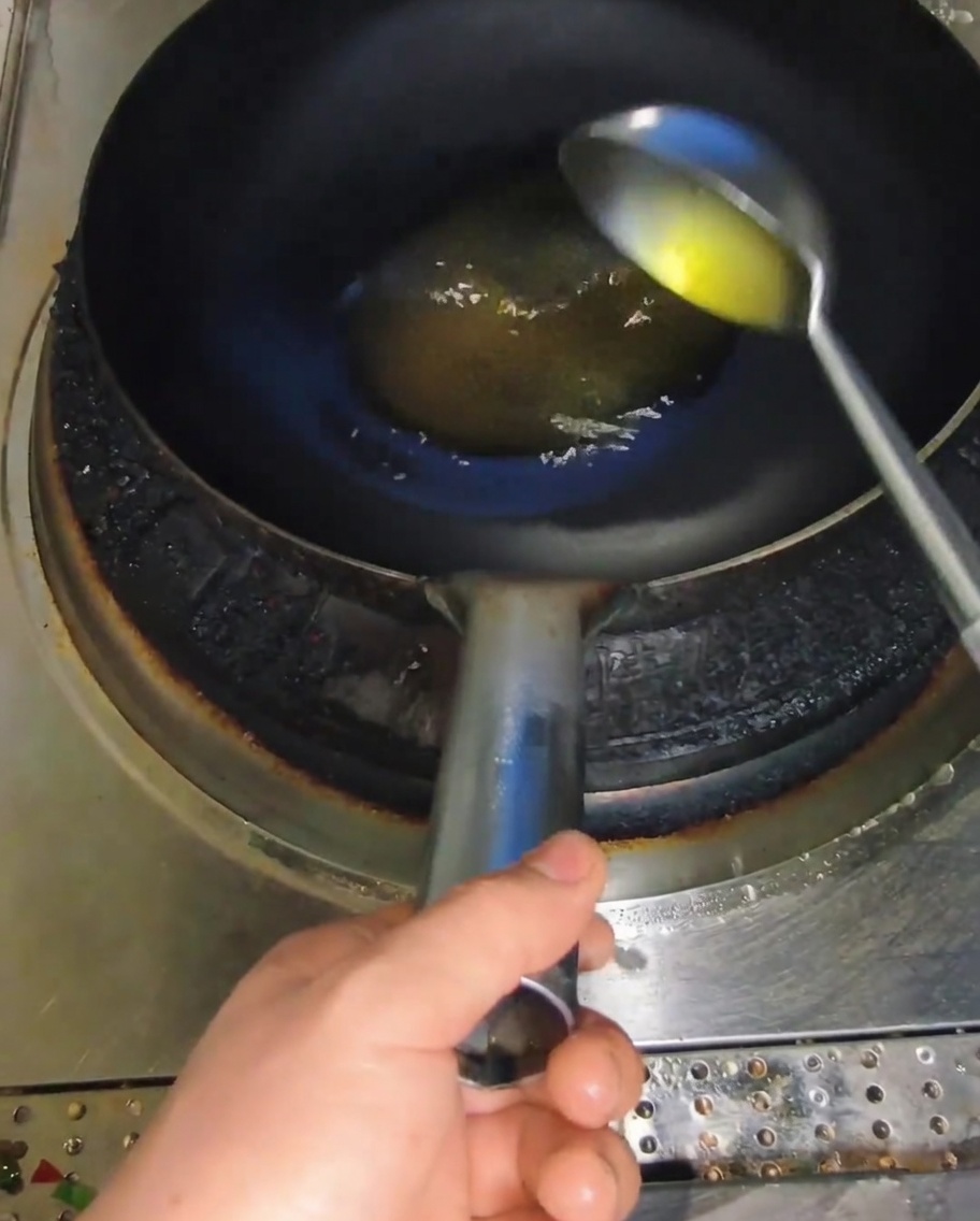 Une louche en métal verse de lhuile de cuisson jaune dans un wok sombre et très chaud.