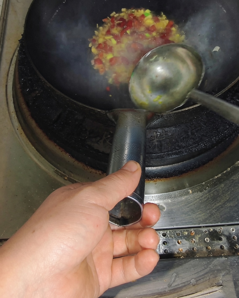 Une louche versant du bouillon clair dans un wok chaud contenant des piments et des oignons verts sautés, produisant de la vapeur.