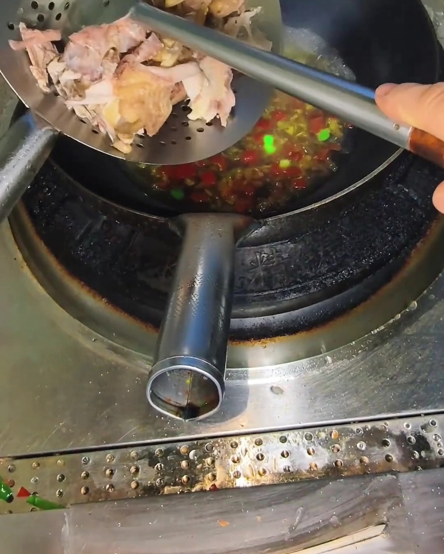 Une écumoire en métal ajoutant des morceaux de poulet cuit et effiloché dans un wok rempli dun bouillon épicé bouillonnant.