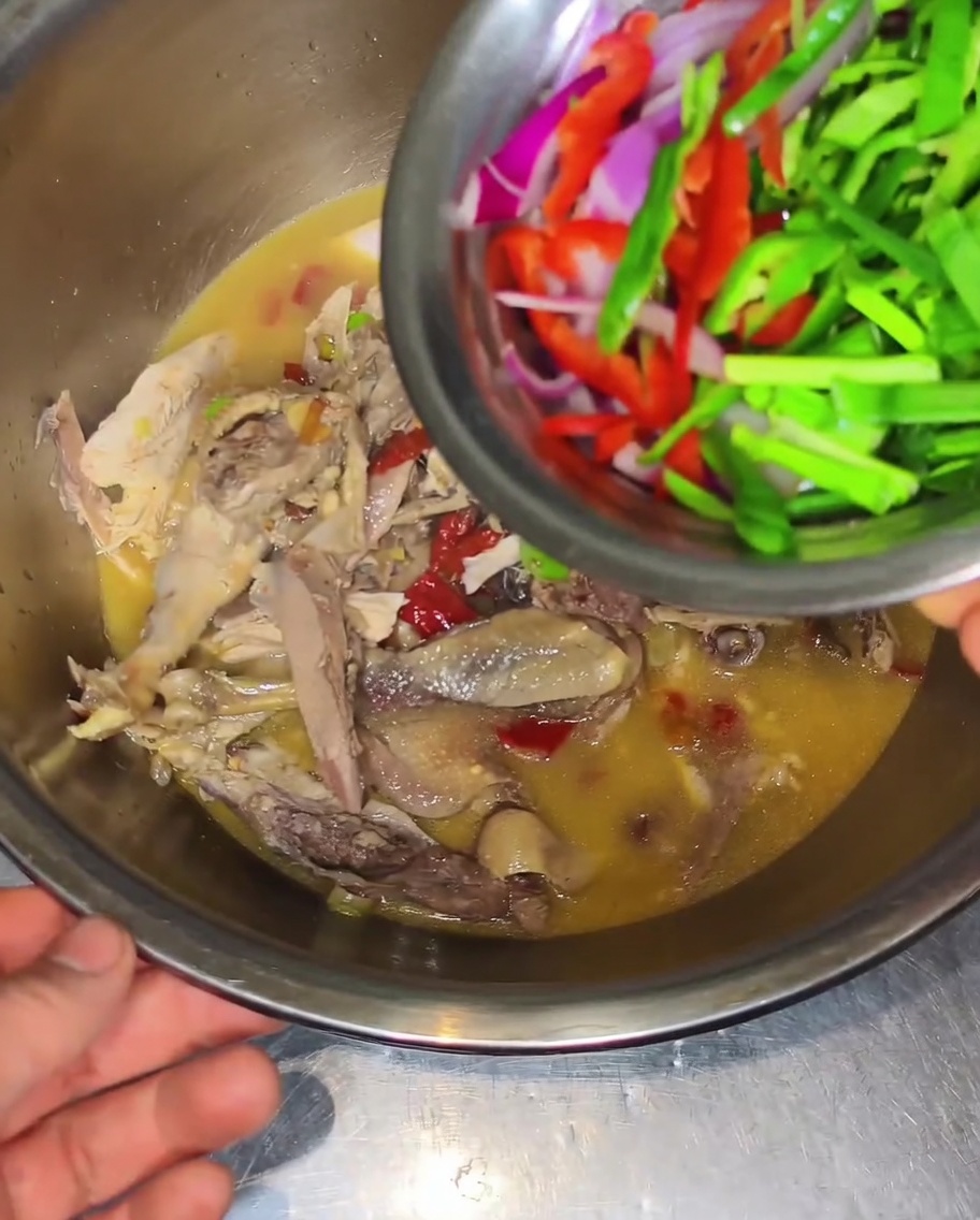 Un petit bol en métal verse de fines tranches doignons rouges, de poivrons verts et de poivrons rouges sur le poulet cuit dans son bouillon au sein dun grand saladier.
