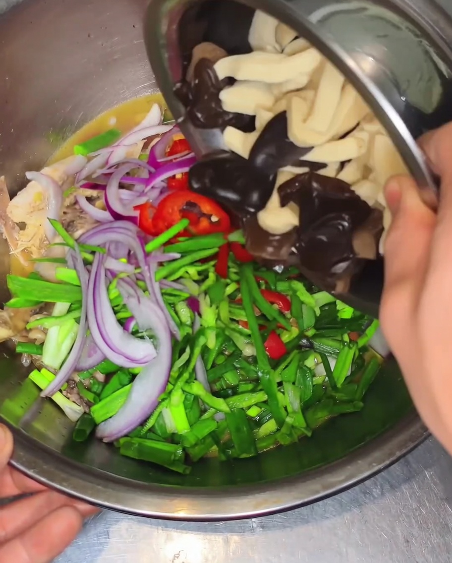 Des mains penchent un petit bol en métal pour ajouter des champignons noirs et dépaisses tranches de peau de tofu dans un grand saladier rempli de poulet, de poivrons et doignons.