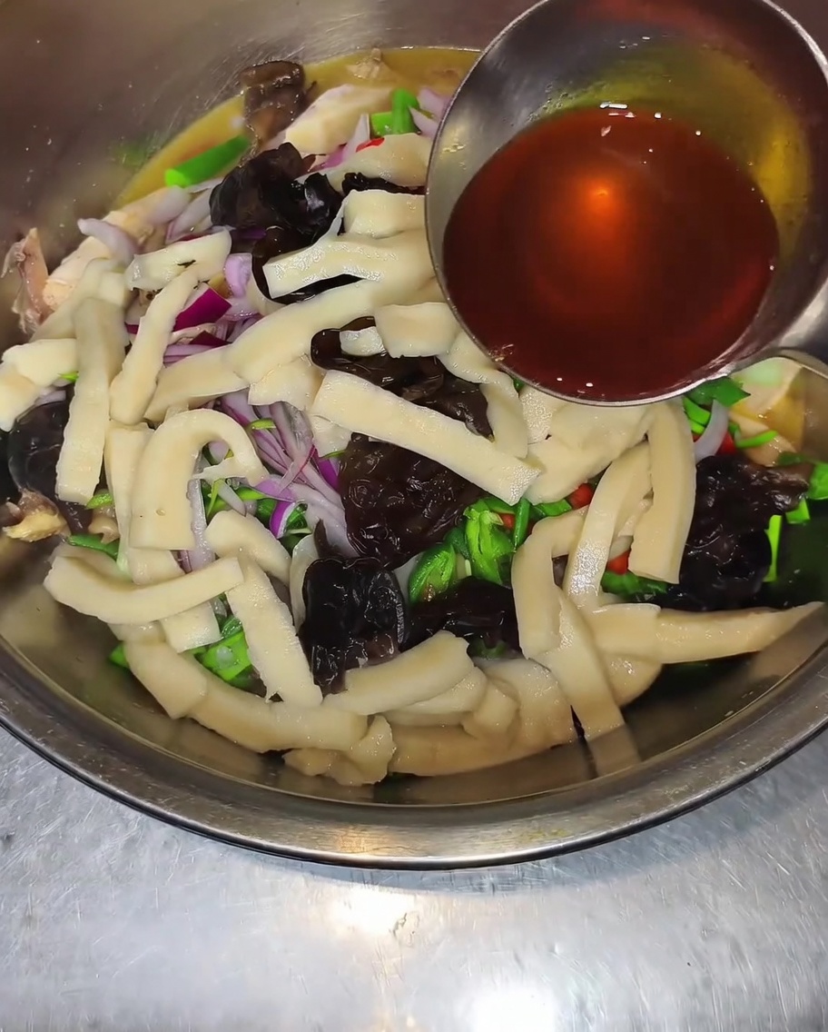 Une louche verse un filet dhuile pimentée sombre et dhuile de poivre sur un bol contenant du poulet, des champignons noirs, des oignons émincés et des poivrons verts.