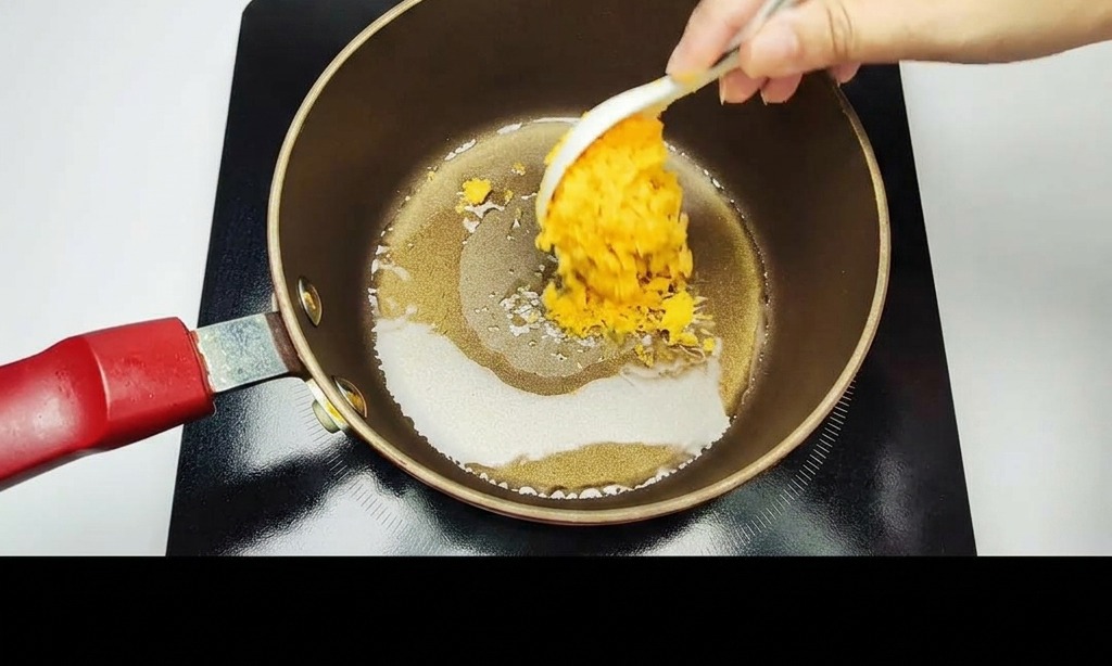 Une main utilisant une cuillère blanche pour faire sauter des jaunes dœufs salés écrasés dans une casserole antiadhésive sombre.