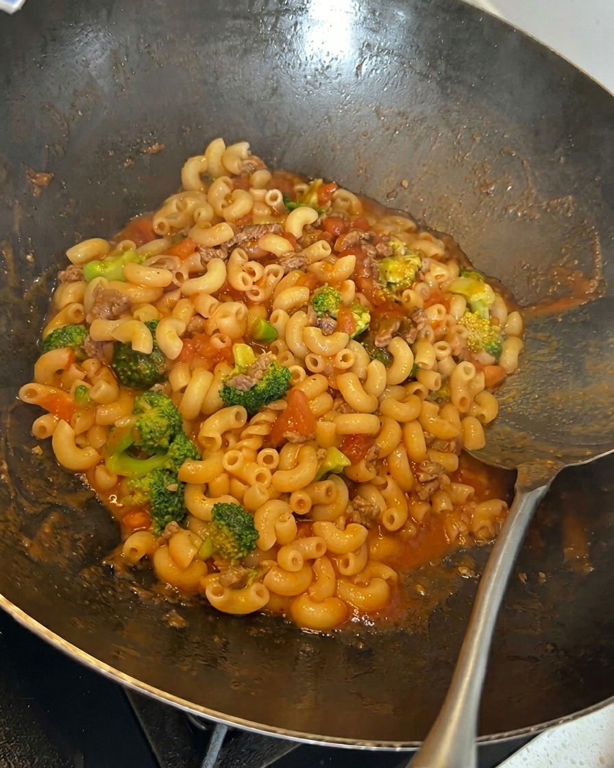 Fromage râpé saupoudré sur un mélange de macaronis, de brocolis et de sauce à la viande et à la tomate dans un wok.