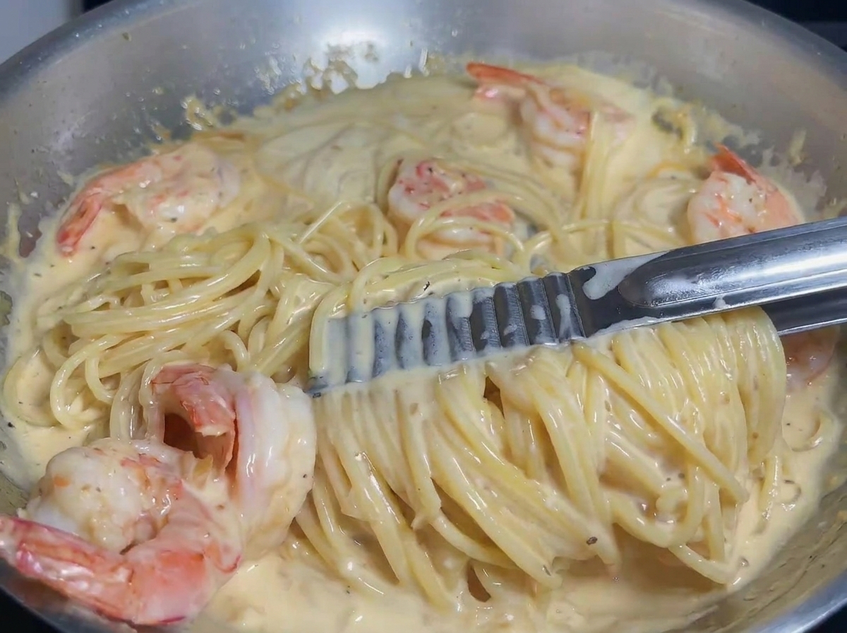 Pâtes et crevettes mélangées à la pince dans une sauce crémeuse pour un enrobage uniforme.
