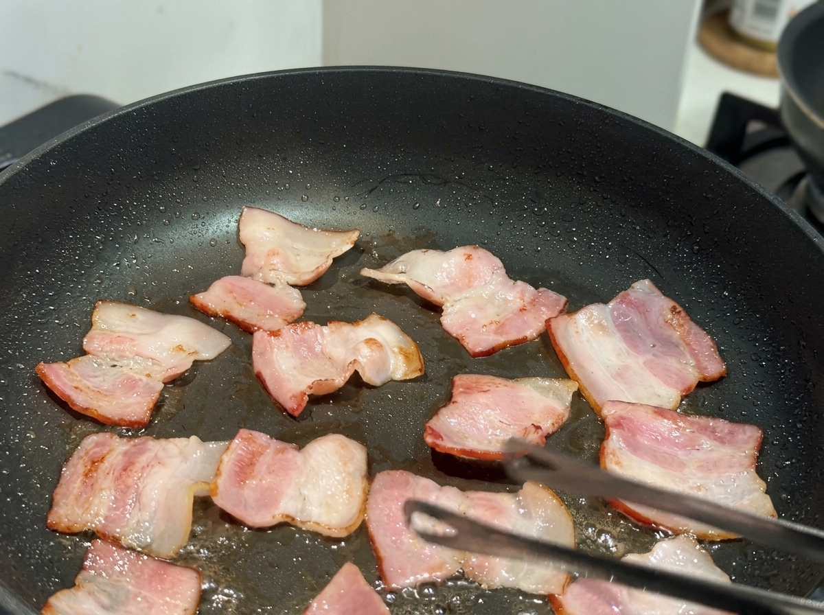 Petits morceaux de bacon plat en train de frire dans une poêle antiadhésive noire avec une pince visible.