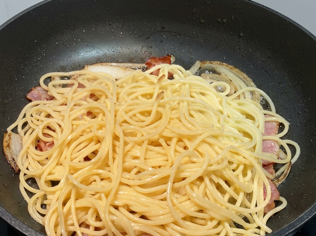Spaghettis cuits posés sur des oignons sautés et des morceaux de bacon dans une poêle noire.