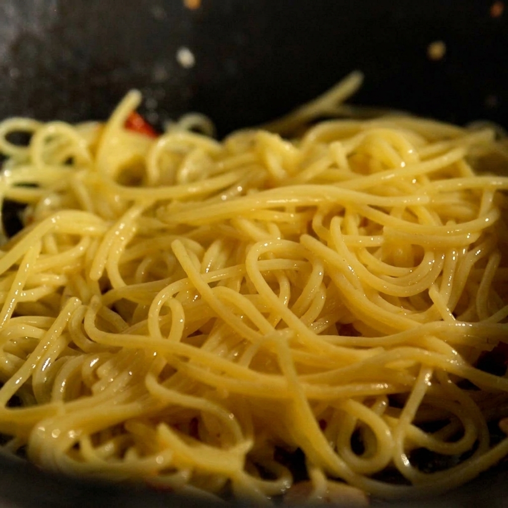 Des nouilles cuites et une noix de beurre ajoutées dans une poêle avec lail et le piment sautés.