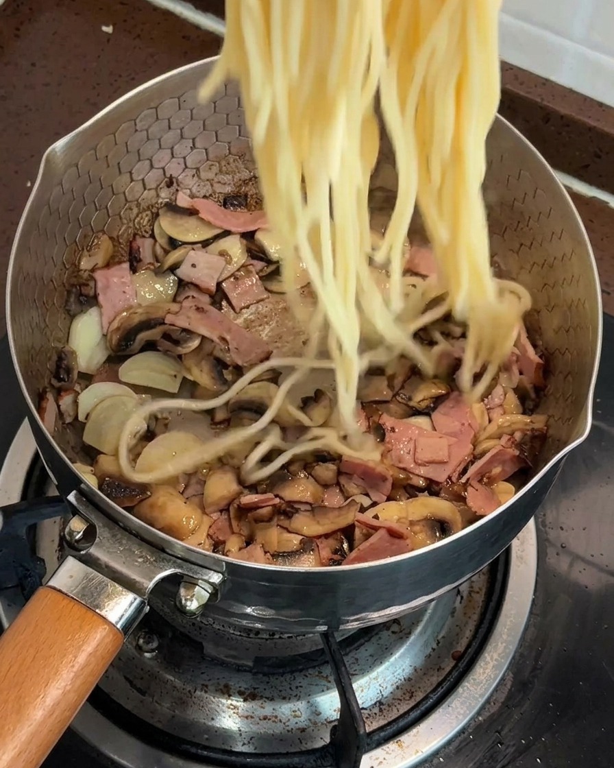 Spaghettis cuites ajoutées directement dans une poêle contenant le mélange de champignons et bacon.