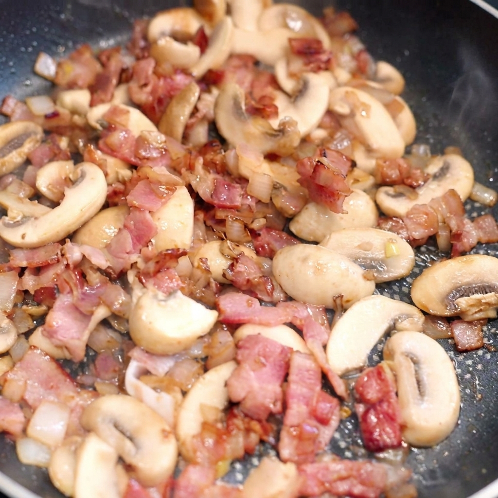 Pâtes larges mélangées dans une poêle avec le bacon doré, les champignons poêlés et les oignons.