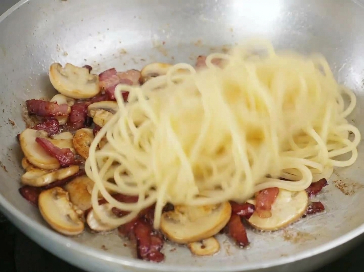 Spaghettis mélangés dans une poêle avec le bacon frit et les champignons émincés.