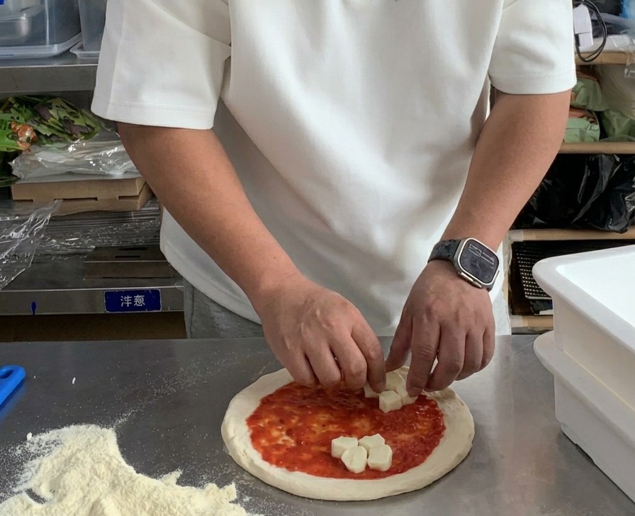 Des mains déposant des cubes de mozzarella fraîche sur une pâte à pizza couverte de sauce tomate.