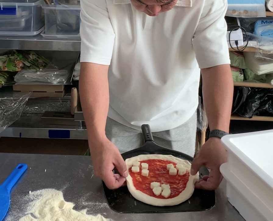 Des mains ajustant les bords ronds dune pizza Margherita non cuite posée sur une pelle à pizza en métal perforé.