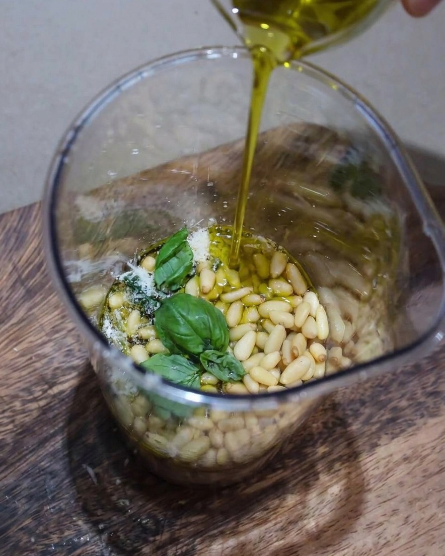 Vue rapprochée des ingrédients du pesto, notamment le basilic frais, les pignons de pin et le parmesan, en cours de mixage dans un bol avec un filet dhuile dolive.