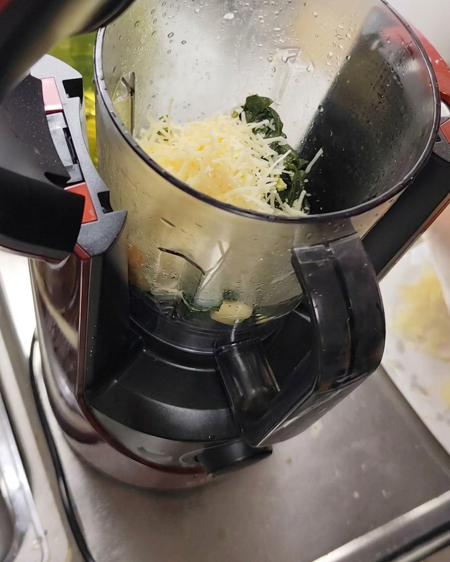 Ingrédients incluant le fromage râpé, lail et les pignons de pin placés dans un robot culinaire avec les feuilles de basilic.