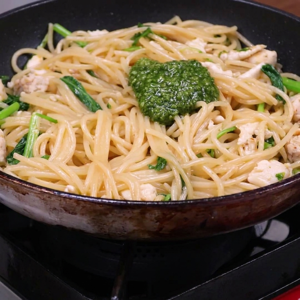 Une cuillerée de pesto vert vif sur un mélange de spaghetti, de poulet et de roquette dans une poêle.