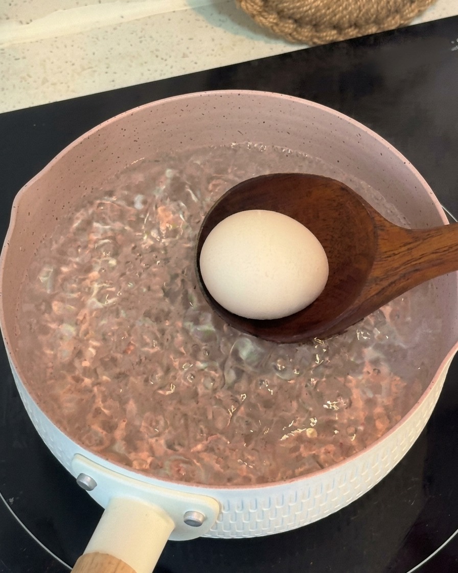 Une louche en bois déposant délicatement un œuf entier dans une casserole rose deau bouillante.