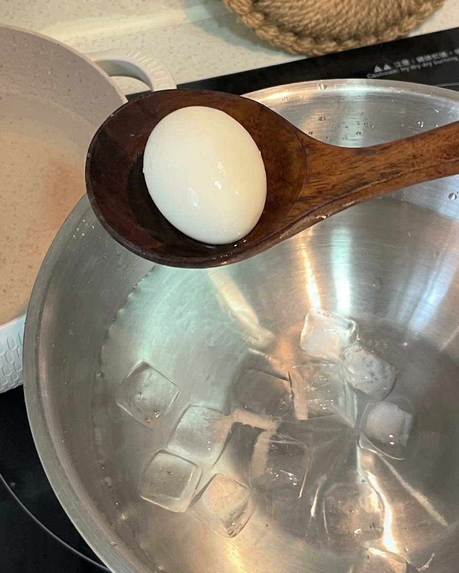Une cuillère en bois transférant un œuf cuit dans un bol en inox rempli deau froide et de glaçons.
