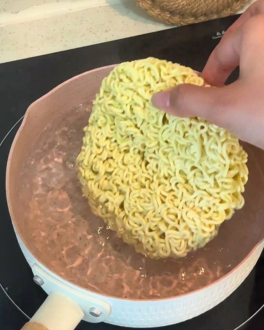 Une main plaçant un bloc sec de nouilles ramen dans une casserole deau bouillante.