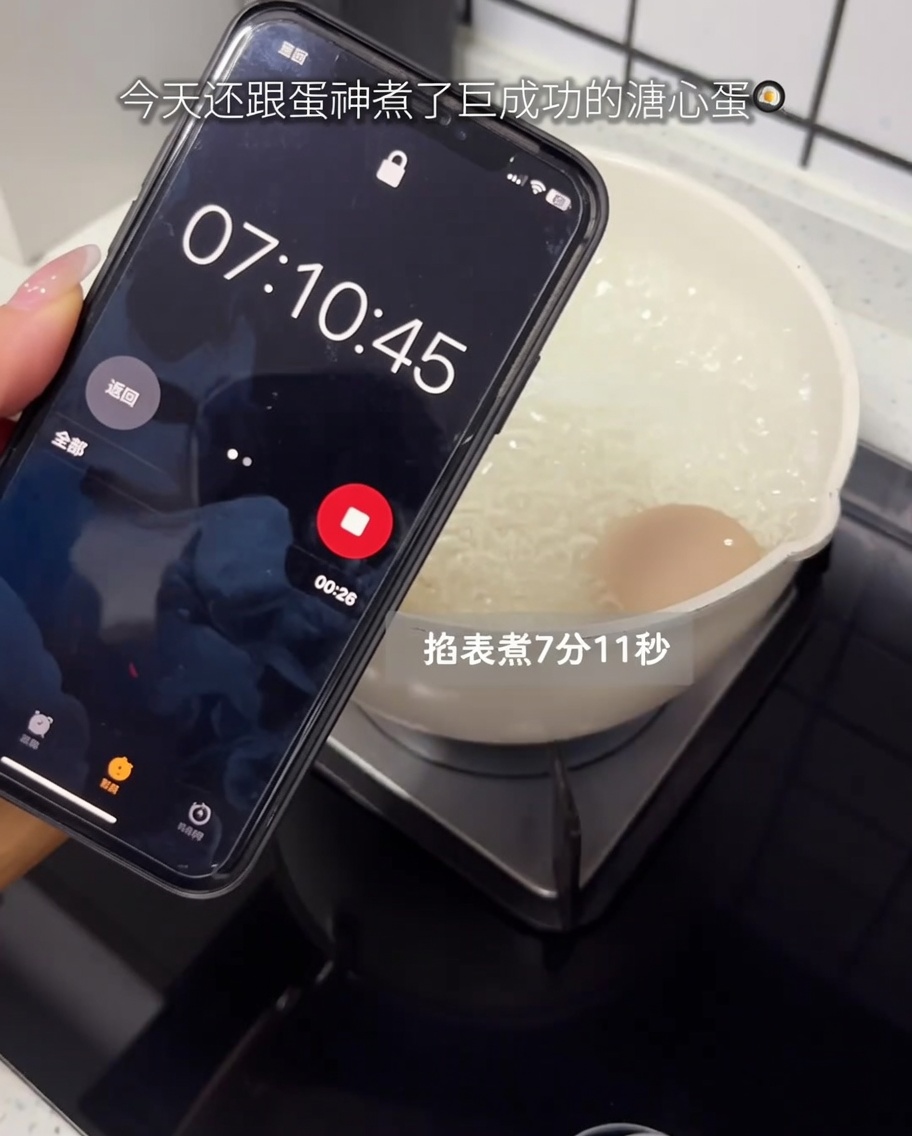 Un œuf cru cuisant dans une casserole sur une plaque de cuisson avec un minuteur de smartphone au premier plan.