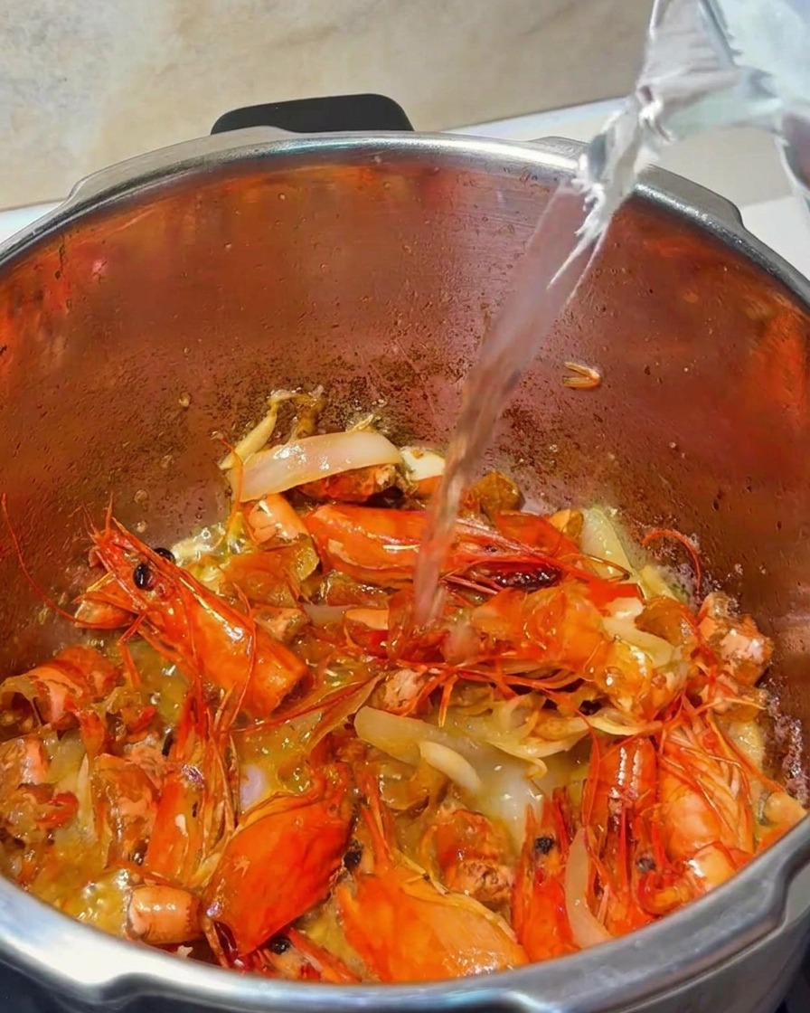 Eau versée depuis un pichet en verre dans une casserole contenant des carapaces de crevettes et des aromates.