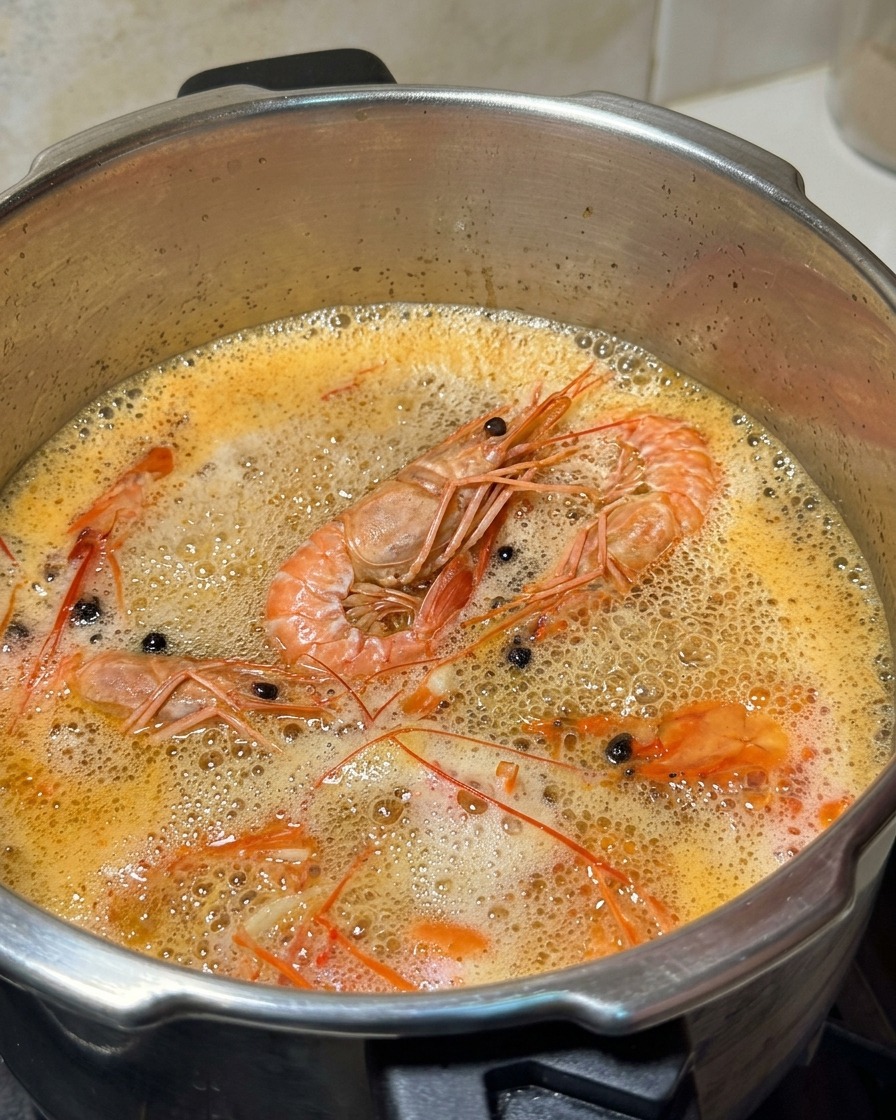 Une louche en métal retirant la mousse blanche à la surface dun bouillon de crevettes orange frémissant.