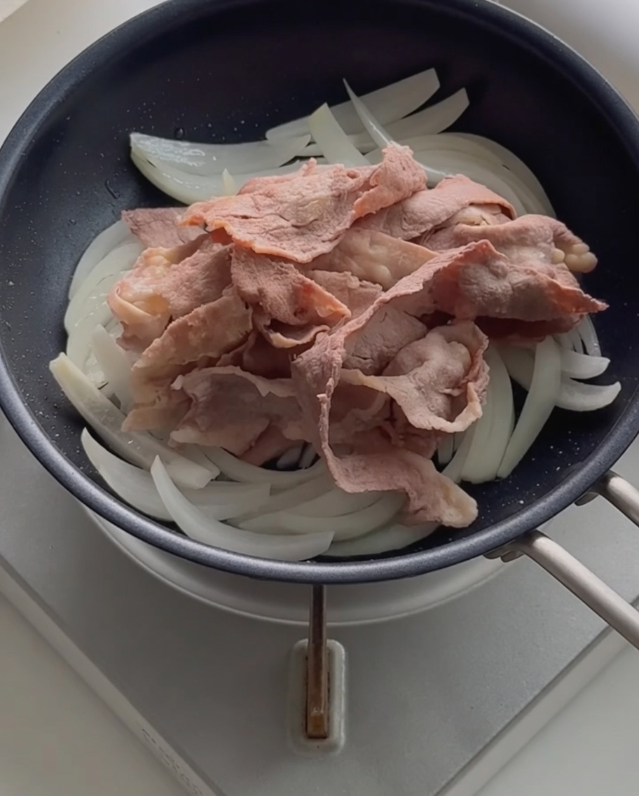 Tranches de bœuf blanchies posées sur des oignons sautés dans une poêle noire.