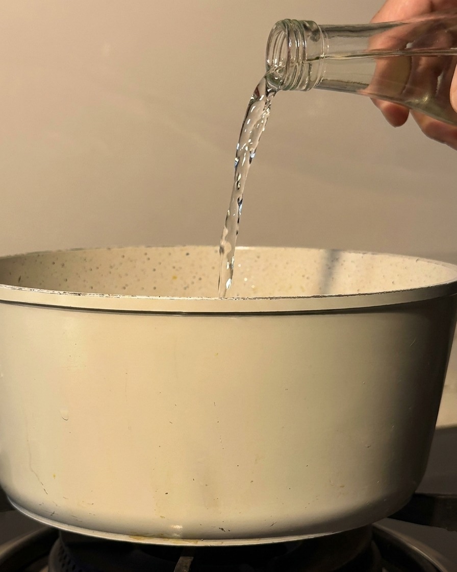 Du saké et du mirin versés depuis une bouteille en verre dans une casserole blanche sur une plaque de cuisson.