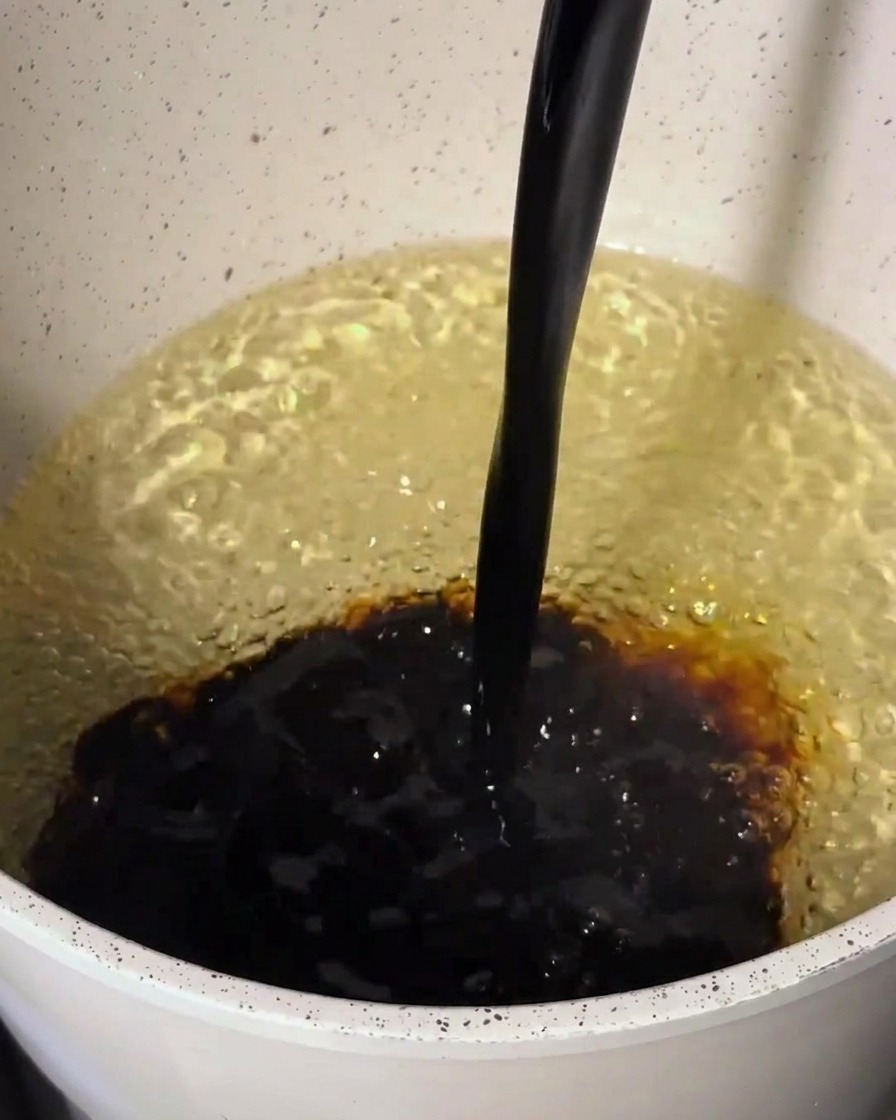 Des ingrédients de sauce brun foncé versés dans une casserole contenant une base plus claire, créant un mélange tourbillonnant.